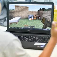 Chłopiec gra w Minecrafta na laptopie, ucząc się jak dodać mody na Aternos. Widok na ekranie gry z łóżkiem, piecem i skrzynią.