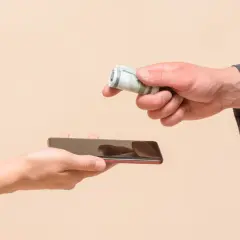 Dwie dłonie, jedna trzyma telefon, druga podaje zwinięte banknoty. Może to być symbol zapłaty za dostęp do informacji, np. jak sprawdzić historię telefonu.