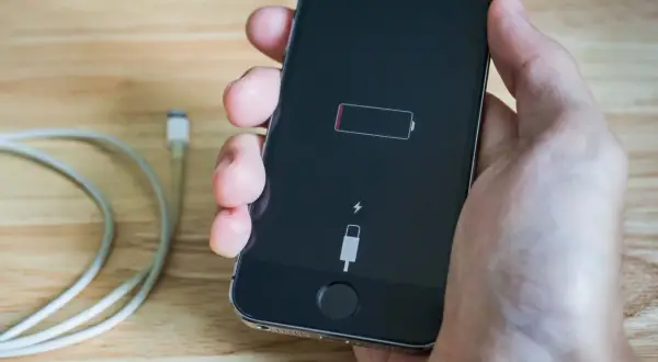 Dłoń trzyma iPhone'a z ikoną pustej baterii i symbolem błyskawicy, co oznacza, że iphone nie ładuje się. Obok leży kabel.
