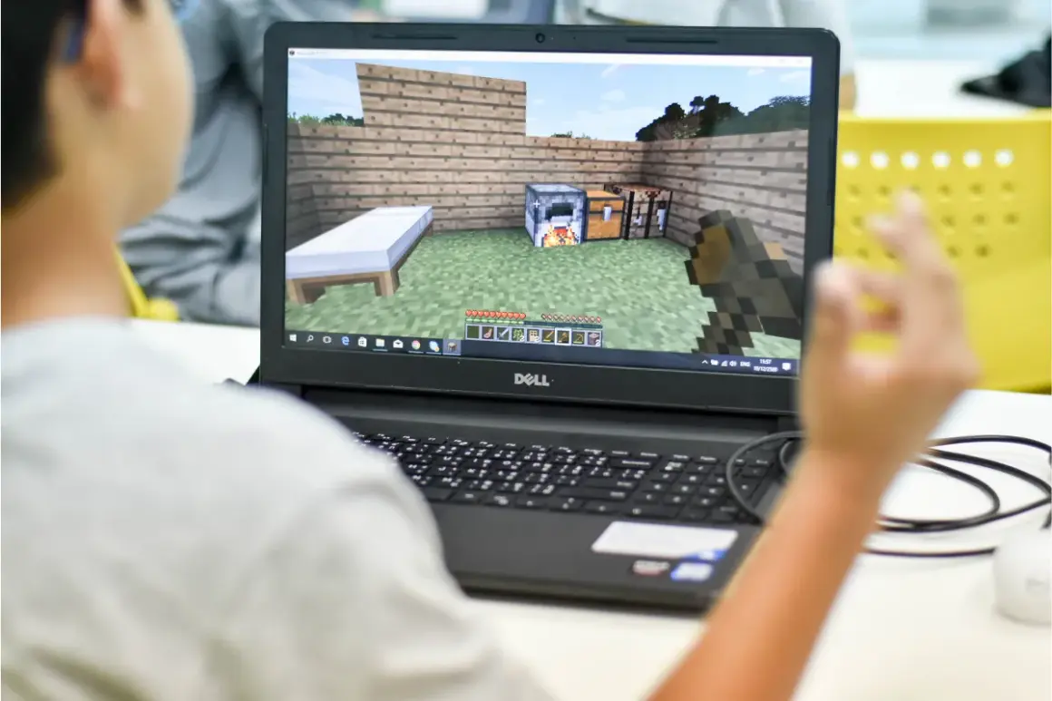 Chłopiec gra w Minecrafta na laptopie, ucząc się jak dodać mody na Aternos. Widok na ekranie gry z łóżkiem, piecem i skrzynią.