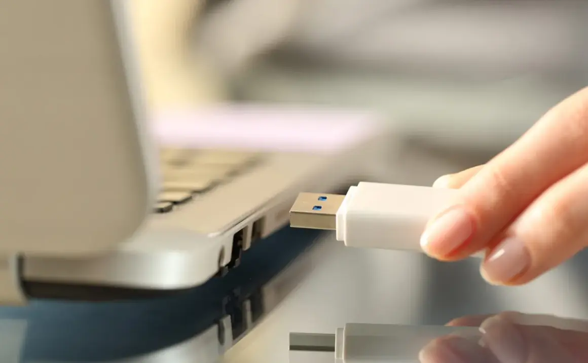 Dłoń wkłada biały pendrive do portu USB laptopa, rozpoczynając nagrywanie danych.