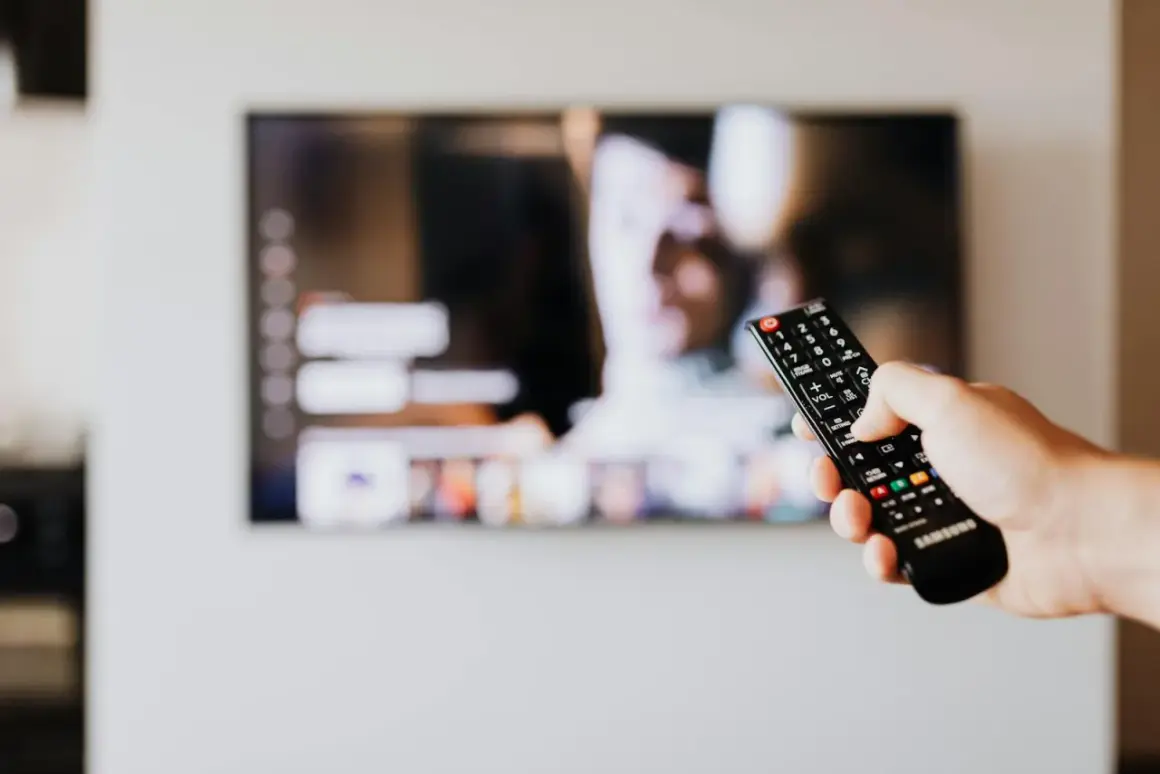 Dłoń trzyma pilot Samsunga, celując w ekran telewizora. Chcesz wiedzieć, jak wyłączyć audiodeskrypcję Samsung?