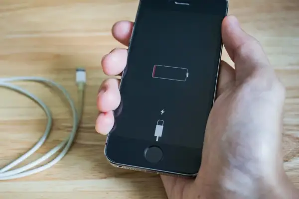 Dłoń trzyma iPhone'a z ikoną pustej baterii i symbolem błyskawicy, co oznacza, że iphone nie ładuje się. Obok leży kabel.