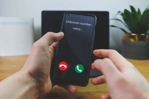 Dłoń trzyma smartfon, na ekranie którego widnieje komunikat "Unknown number calling...". Numer prywatny dzwoni, a użytkownik zastanawia się, czy odebrać.