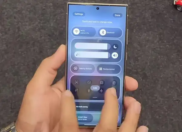 Dłoń steruje smartfonem z interfejsem One UI, pokazując panel szybkich ustawień z Wi-Fi, Bluetooth i innymi opcjami.